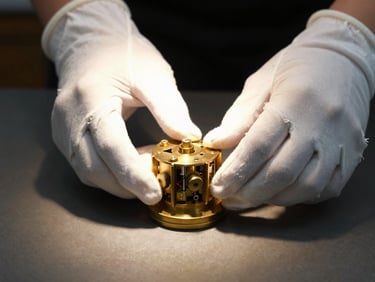 Close-up of hands in white cotton curatorial gloves carefully adjusting an intricate brass mechanical cylinder, single-source warm spotlighting casting deep shadows on a dark table.