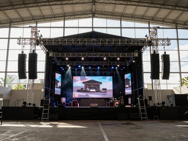 Wide environmental shot from the back of house looking toward a fully-rigged concert stage in a large Monterrey venue — speaker arrays flanking both sides, moving-head lights pointing at the stage, LED screen panels displaying live content at center, the room ready before doors open, crisp daylight from venue windows cutting across the rigging, no performers visible