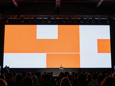 Wide environmental shot from control position at the back of a live Monterrey event — a large LED video wall at the front of the venue displaying vivid orange and white presentation content, the crowd visible in silhouette in the foreground, the screen bright and sharp against the dark venue ceiling, technical readiness evident, no performer close-ups