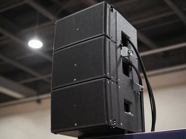 Close-up of a professional speaker array column mounted in position inside a large venue, cables neatly routed, ambient venue lighting visible in background, shot from slightly below looking up to show scale and readiness, no people