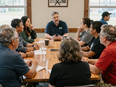 A warm, natural light photograph of a community meeting in Oregon, Ohio, showing staff members talking face-to-face with local residents around a wooden table, unposed documentary style.
