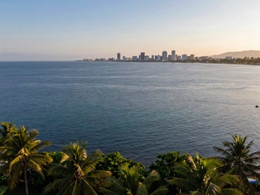 Elevated wide-angle photograph of the Davao Gulf during late afternoon, showing calm deep blue water with the distant outline of Davao City under a soft golden haze.