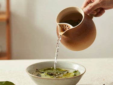 Hands gently pouring hot water from a clay kettle into a ceramic bowl with loose green leaves