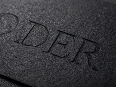 Macro crop of debossed serif typography on heavy textured black cotton cardstock, hard directional key light, deep shadows.