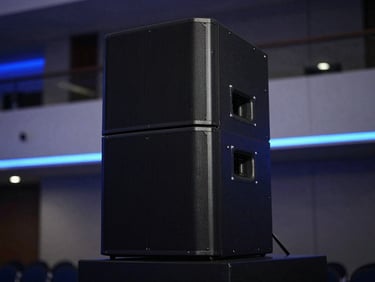 Sleek matte-black professional speaker array standing in a modern architectural event hall, low-key ambient lighting with electric blue neon accents, shallow depth of field focusing on the clean speaker chassis, no cable clutter.
