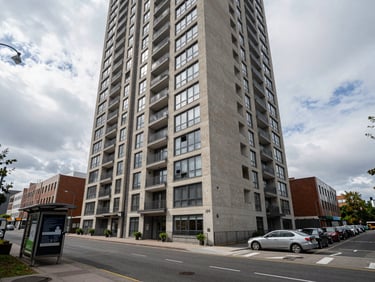 Wide exterior of a compact urban apartment tower on a busy street, overcast daylight, footpath and nearby transit stop visible in foreground for location context, no models