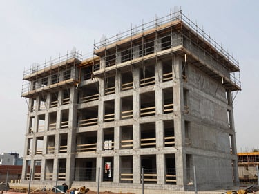 Wide exterior shot of a six-storey residential building mid-construction, scaffolding on upper two floors, concrete structure visible, overcast natural daylight, site boundary fencing in foreground