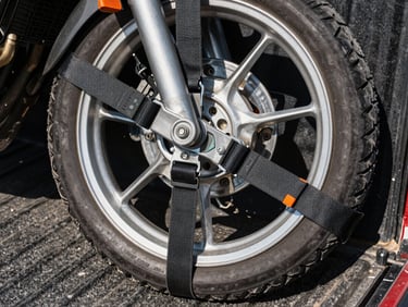 Close-up overhead shot of heavy-duty ratchet tie-down straps securing a motorcycle wheel to a pickup truck bed, strong midday sun casting sharp shadows on orange-tipped equipment handles, asphalt texture visible at edges