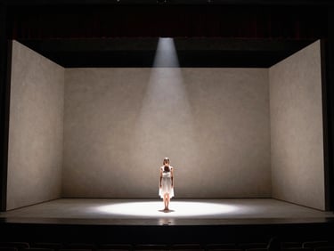 Wide production still of a minimalist plaster theatre stage, a single female performer standing in a dramatic shaft of light, deep shadows.