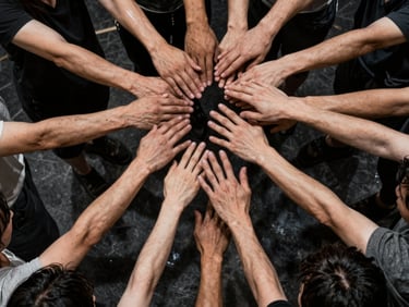 High-contrast rehearsal photo of an ensemble of actors in a tight physical composition, hands reaching upward, raw charcoal tones.