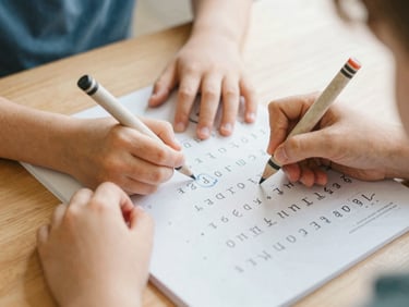 Children's hands holding a thick crayon, tracing letters on a printable workbook page on a wooden table, soft morning light