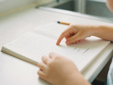 Close-up of a child's hands carefully turning a page of a printed workbook on a quiet kitchen table, warm morning light