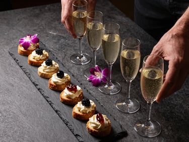 Overhead shot of a cocktail reception spread — hand-passed canapés on a black slate board, champagne coupes arranged in a diagonal line, a single orchid sprig accent, dark stone surface, studio strobe balanced with warm ambient light