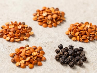 Overhead macro photograph of vibrant orange achiote seeds and whole black peppercorns arranged in clean geometric piles on a light textured stone surface, soft natural side lighting.