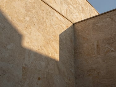 Low-angle golden hour shot of raw, textured plaster and honed travertine stone walls, deep shadows, minimalist architectural lines, no people