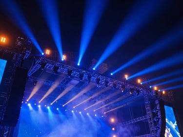Low-angle heroic shot of massive concert stage rigging under vibrant cobalt blue and safety orange lights, haze in the air, technical precision, sharp focus.