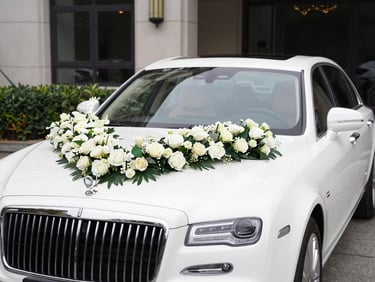 A luxury white sedan wedding car decorated with fresh flowers on the hood, parked in front of an elegant venue, bright daylight