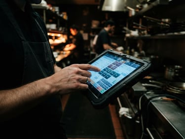 Close-up of a rugged Android tablet displaying a native ordering UI, held by a server's hand mid-service in a dimly lit, high-contrast kitchen, cool screen glow, rich dark tones, 35mm lens.