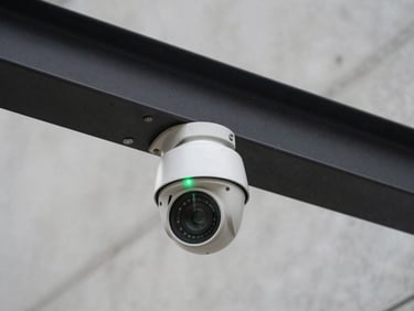 Straight-on elevation of an industrial security camera enclosure mounted on a matte black metal beam, native green LED status light active, clean concrete background.