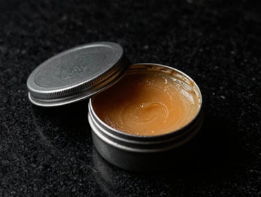 A premium metal tin of hair pomade open on a black marble surface, rich texture of the wax visible, dramatic low-key lighting.