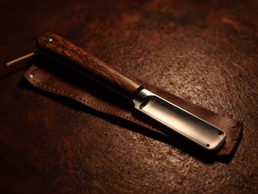 A classic straight razor with a dark wood handle resting on a leather strop, steel blade catching a sharp amber highlight, deep shadows.