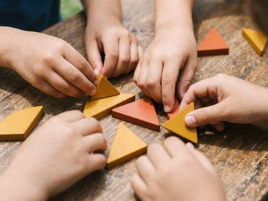 Close-up of children's hands arranging geometric wooden shapes in warm mustard and terracotta colors on a rustic table, bright natural daylight, shallow depth of field.