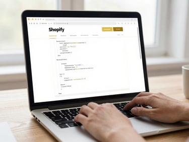 A close-up of hands typing code on a laptop screen displaying a clean, modern Shopify store dashboard, bright editorial style, warm gold accents