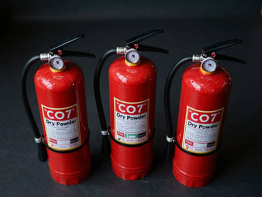Overhead view of three fire extinguishers — CO2, dry powder, and wet chemical — arranged on a dark floor, cool studio strobe from above, labels and pressure gauges in focus