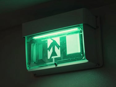 Close-up of an emergency exit luminaire mounted on a dark wall, green LED diffuser panel glowing sharply, cool studio light on the housing body, architectural context visible behind
