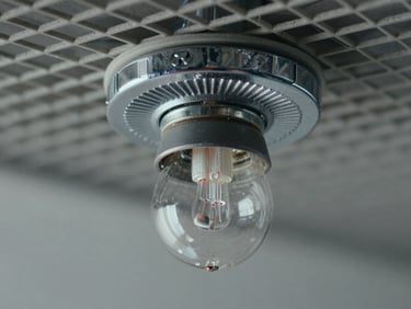 Extreme close-up of a pendant sprinkler head mounted in a suspended ceiling grid, chrome deflector plate and glass bulb in sharp focus, cool diffuse studio light, neutral background