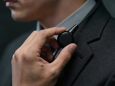 Close-up of a security agent's hand adjusting a communications earpiece, sharp focus, dark tailored suit sleeve, low-key lighting