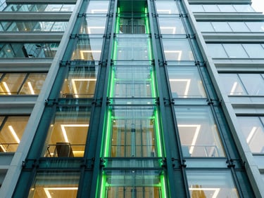 Symmetrical architectural shot of a custom inclined glass elevator on an ultra-modern building facade, bright daylight mixed with neon green accent lights, wide-angle lens