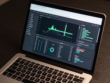 Close-up overhead view of a laptop screen displaying a complex analytics dashboard interface in Figma, multiple data panels visible, even studio lighting, dark desk surface visible at edges