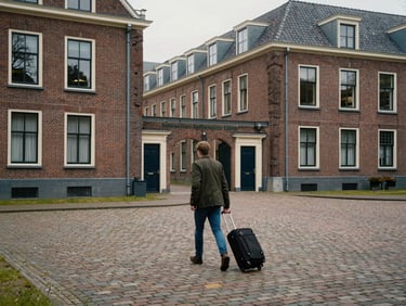 Wide environmental shot of a Dutch university campus courtyard in overcast light, a student pulling a rolling suitcase across cobblestones toward a brick building entrance, seen from behind at mid-distance