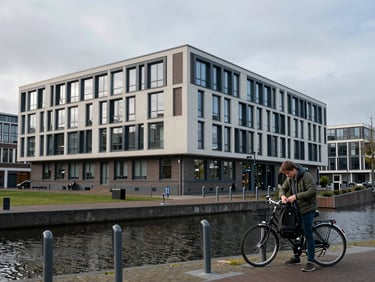 Environmental shot of a Dutch university campus — a modern low-rise faculty building beside a canal, a student at a bicycle rack removing a backpack, flat grey sky, cool diffuse daylight, calm urban academic context