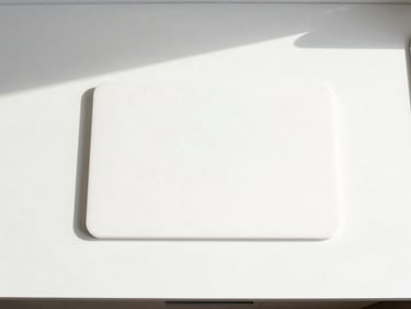 Overhead flat-lay of minimalist kitchen utensils and a sleek cutting board on a white counter, bright natural daylight.