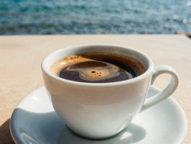 Rich dark espresso shot with thick crema in a white ceramic cup on a sun-drenched outdoor table by the sea