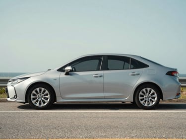 Three-quarter exterior shot of a silver Toyota Corolla sedan parked on a Cayman roadside in natural daylight, full vehicle profile visible, slight road texture in foreground