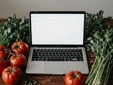 Overhead shot of a laptop displaying a social media calendar, surrounded by fresh seasonal produce like tomatoes and herbs, in soft Scandinavian daylight, matte finish.