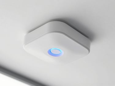 Close-up of a sleek white ceiling-mounted UniFi access point with a soft blue circular light glowing in a bright modern office, crisp industrial photography
