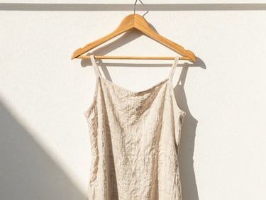 Bright editorial shot of a linen dress hanging on a wooden hanger against a clean textured wall, soft shadows
