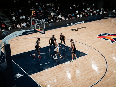 Overhead wide shot of a basketball court mid-game, players clustered under the basket, venue lighting casting hard shadows, sponsor banners visible along the sideline