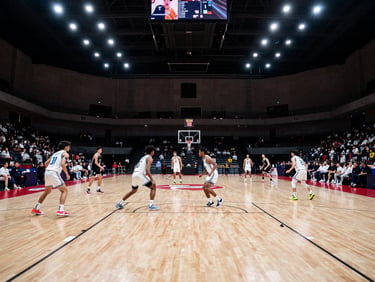 Indoor basketball court wide shot, two teams in a fast break, overhead gymnasium lighting, scoreboard visible in the upper frame, crowd seated courtside