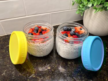 Two glass jars of chia pudding topped with fresh berries with colorful lids on a kitchen counter.