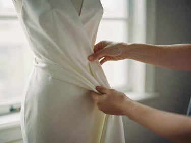 Warm editorial close-up of a designer's hands draping heavy silk fabric on a mannequin, soft side-lit window daylight, desaturated charcoal and ivory tones, shallow depth of field, 35mm