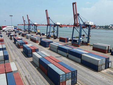 Aerial drone shot of a busy Indonesian port terminal at midday, rows of shipping containers in navy and grey stacked at loading bays, container cranes visible against a clear sky, no people in foreground, wide environmental framing showing port scale and operational order