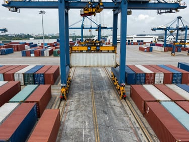 Overhead shot of shipping containers being loaded at a port loading bay, crane visible at top frame, containers in neat rows, cool overcast daylight, industrial port scale