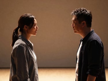 Two theatre actors face-to-face in intense dialogue on a bare stage, dramatic side-lighting in warm gold, deep shadows framing their expressions