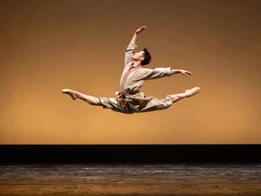 Dancer in full extension mid-air, traditional cloth billowing, golden-hour light flooding from stage left, dark stage floor below reflecting amber glow