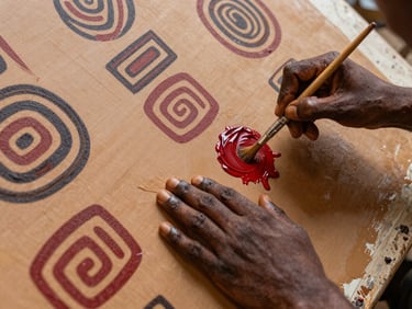 Overhead composition of a painter's hands applying ochre and deep red pigment on a large canvas, natural north-facing daylight, fine brushwork visible, fragments of Adinkra geometric motifs emerging on the surface